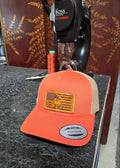 An orange and tan trucker hat with a rectangular American flag patch featuring a deer silhouette, resting on a dark countertop in front of a sewing machine.