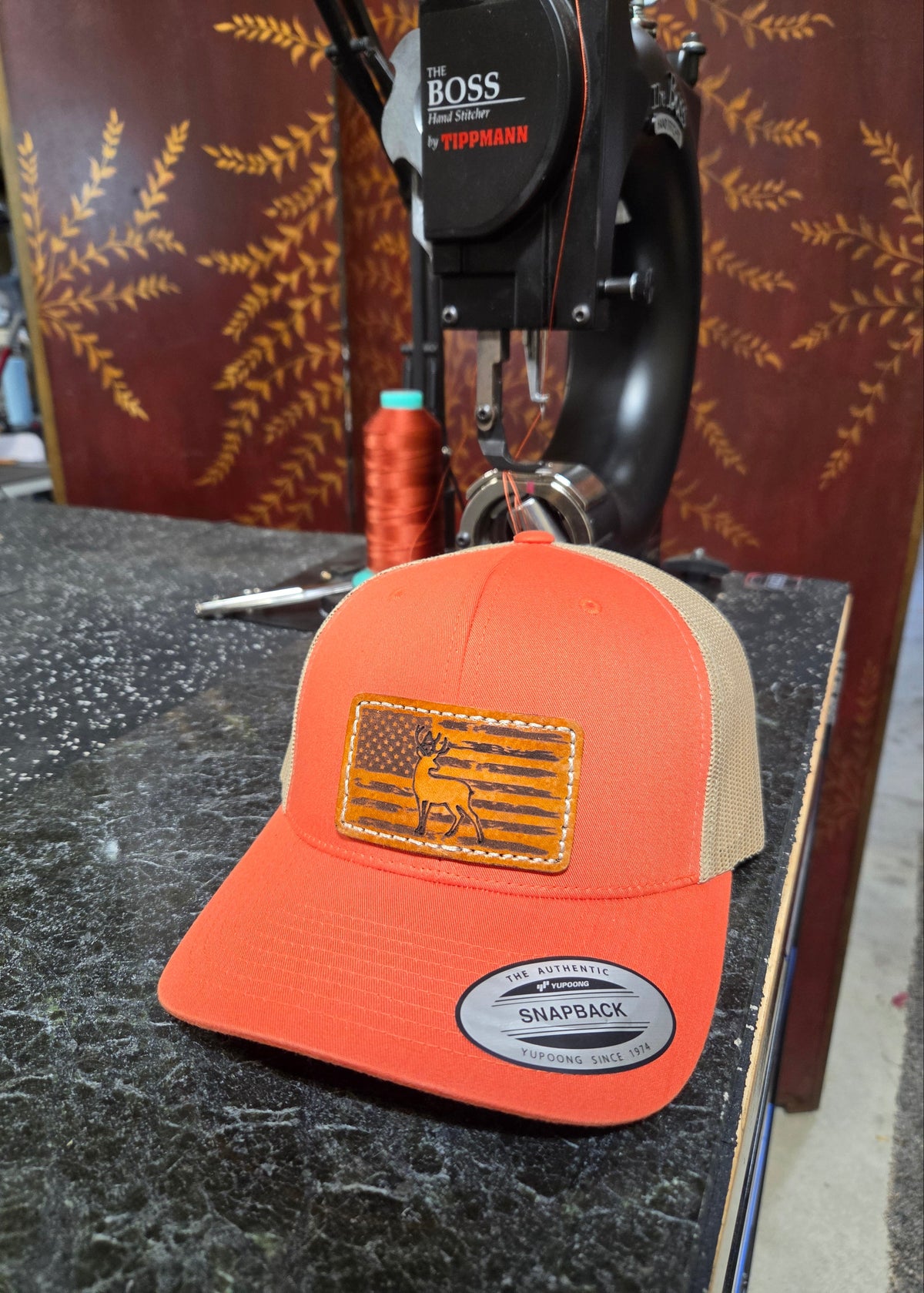 An orange and tan trucker hat with a rectangular American flag patch featuring a deer silhouette, resting on a dark countertop in front of a sewing machine.